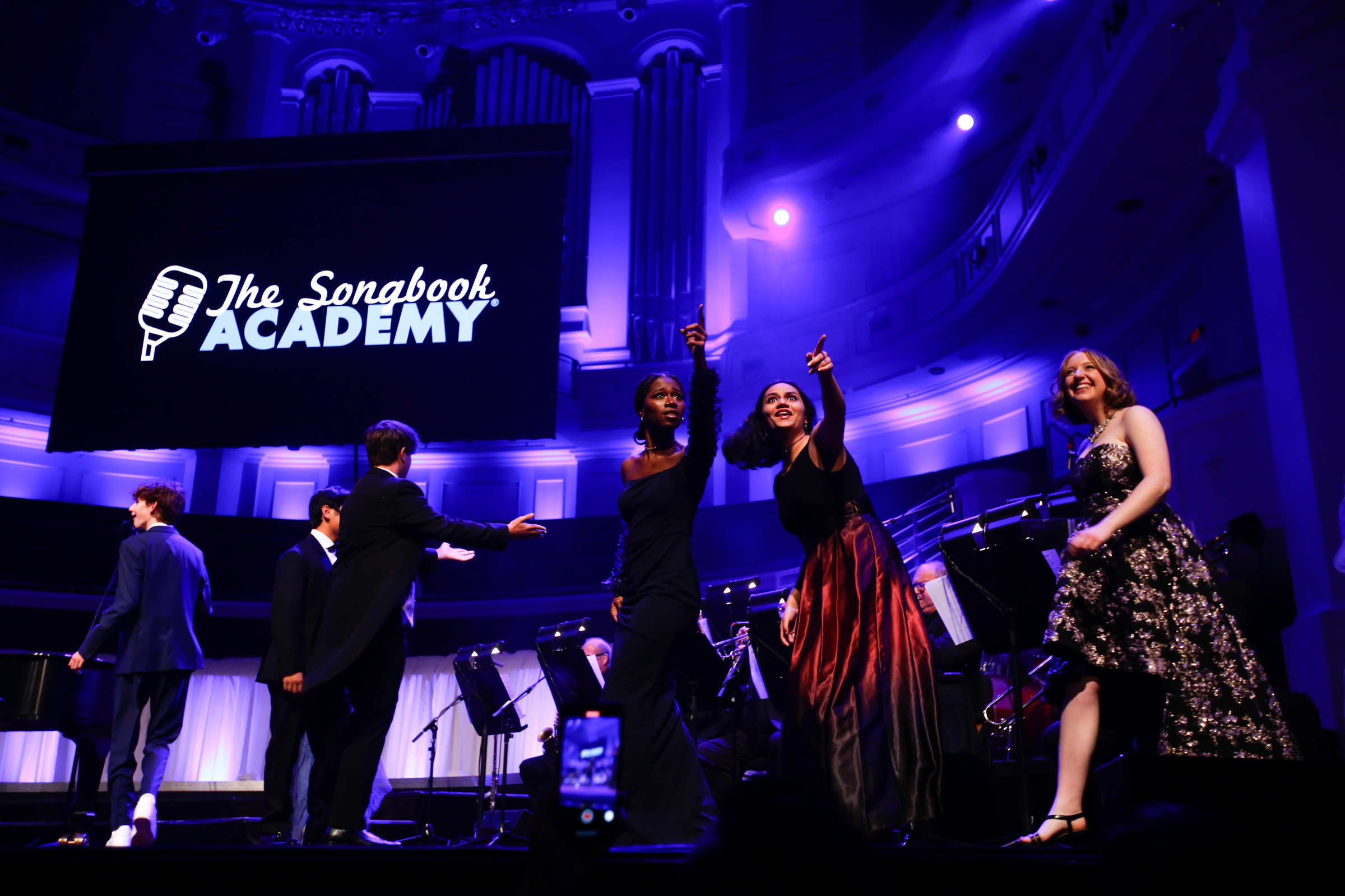 Teen singers in evening wear point at the audience as they walk onstage during the annual Songbook Academy final concert.