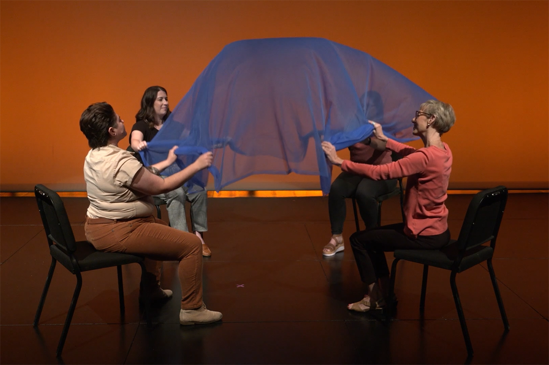 Seated in chairs on a stage, four women demonstrate a relaxation exercise using a large blue scarf.