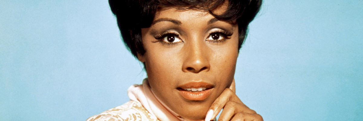 Diahann Carrol looks straight ahead at the camera and smiles demurly. Her hand rests on her jaw and her eyebrow is cocked.