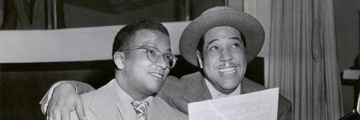 Duke Ellington wearing a hat and holding sheet music, puts his arms around the bespectacled Billy Strayhorn.
