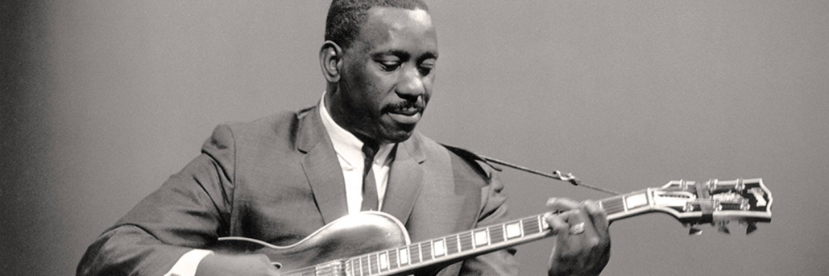 Wes Montgomery is in a nice suit and looks down at an electric guitar he plays. He has tight cropped hair and a moustache.