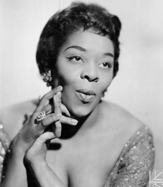 African American singer, Dinah Washington purses her lips and looks to the side with her hands clasped together.