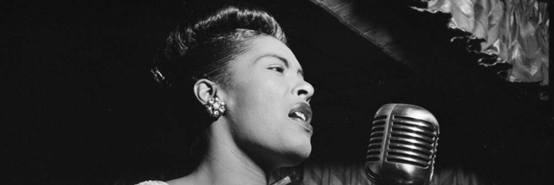 African-American vocalist, Billie Holiday sings in an old-fashioned microphone on a darkened stage.