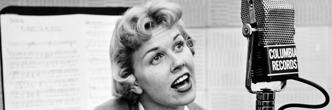 Blonde singer, Doris Day, sings into a Columbia Records microphone. Behind her is sheet music.