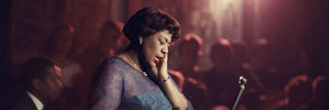 Ella Fitzgerald, a Black woman in a purple dress, hold her ear, closers her eyes and sings in a darkened club.