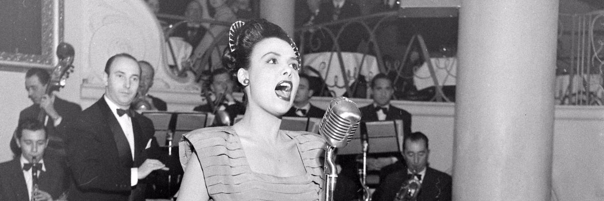 Lena Horne, a light skinned and stunning Black woman, sings into a microphone at a club. A full band is behind her.