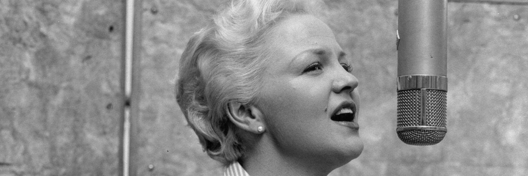 A blonde Peggy Lee cocks her head up toward a microphone and sings.