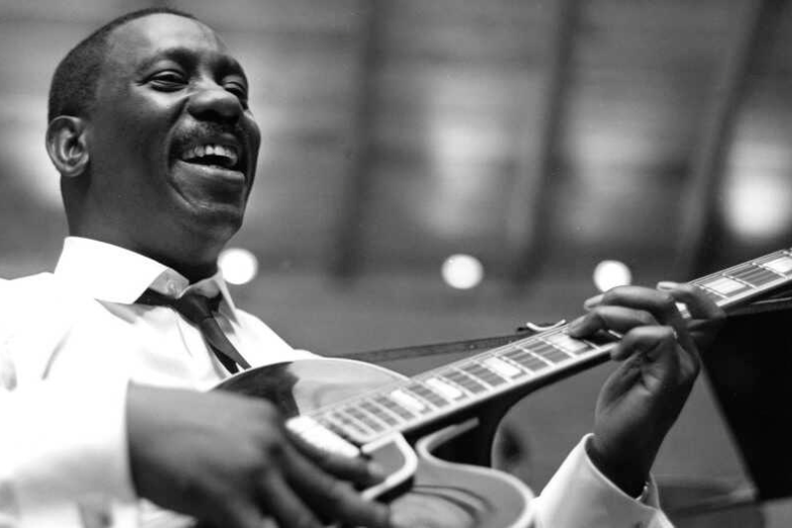 Wes Montgomery, a smiling Black man, wears a nice button down shirt plays a guitar.