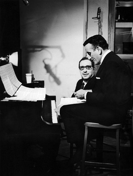 Harold Arlen sits at a piano covered in sheet music and looks up at a young Tony Bennett who looks at sheet music. They are in a darkened studio apartment.