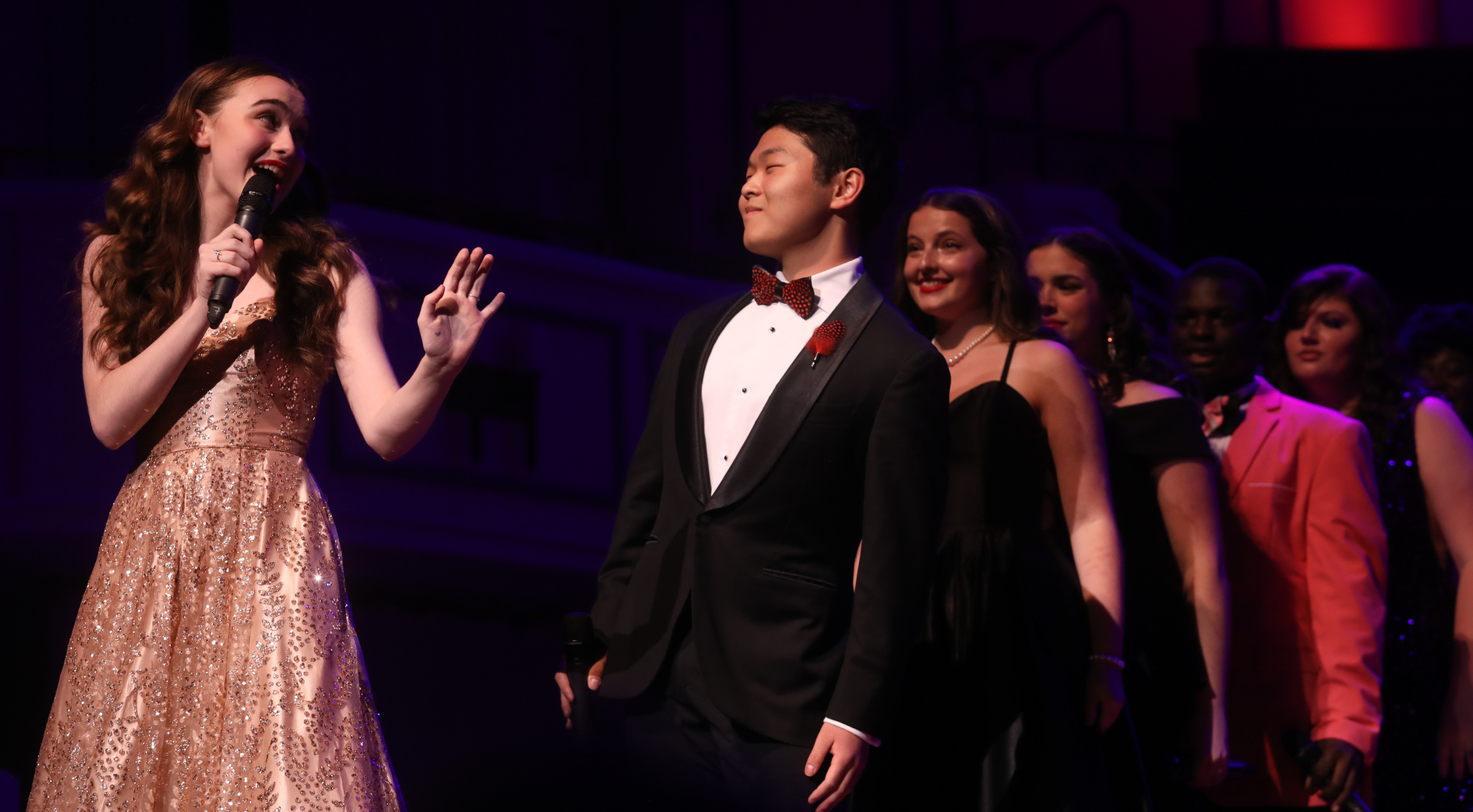 A young woman with wavy hair and a champagne dress holds a microphone, looks back and sings to a group of high school singers in formal wear.