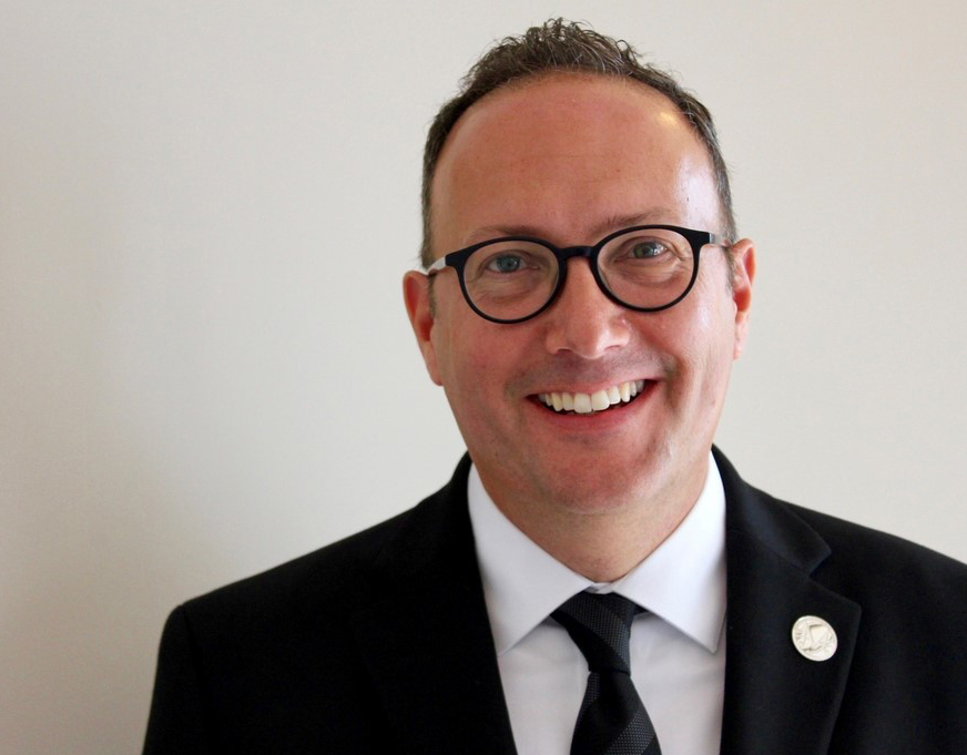 A man wearing a dark suit and eyeglasses smiles.