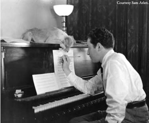 Composer Harold Arlen sits at a piano and writes on a piece of sheet music. A small dog is drapped over the piano.