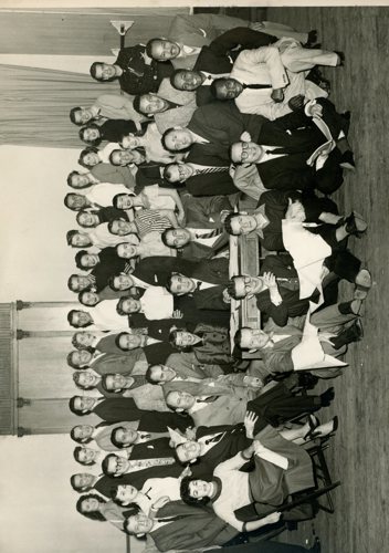 Sepia image of a large group of men and women dressed semi-formally in suits and ties, skirts, sweaters, all smiling at the camera.