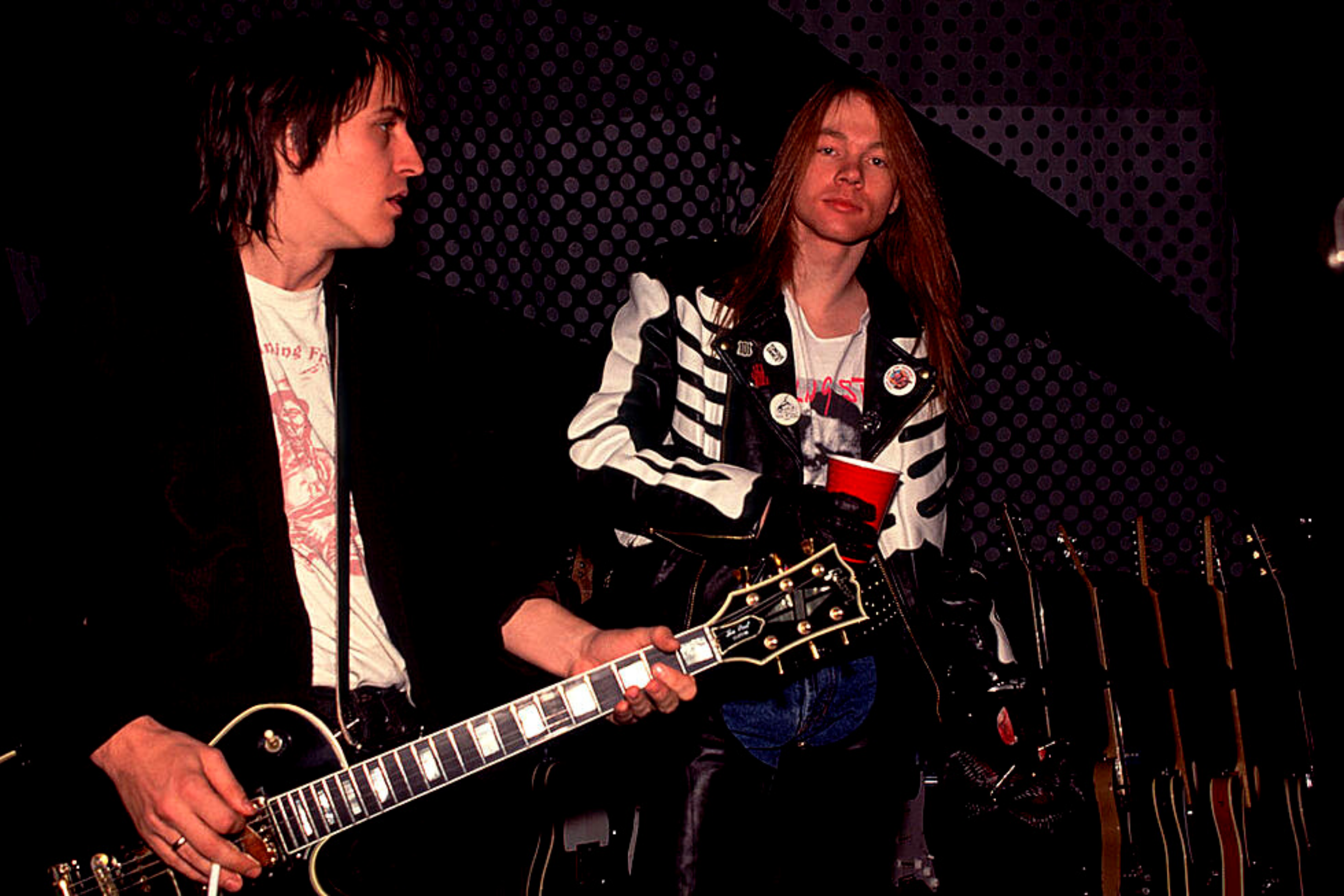 Izzy Stradlin holds a cigarette and plays his guitar while Axl Rose stares beyond the camera.