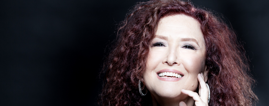 Melissa Manchester with wavy red hair smiles with her hand touching her jaw.
