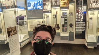 Chelsea wears a black face mask, round glasses and poses in front of the jazz section of the Musical Crossroads exhibit.