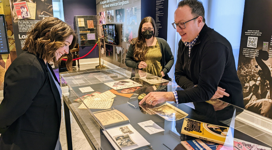 Chris points to a scrapbook in an exhibit case. Renee and Emily bend over and smile at the object.