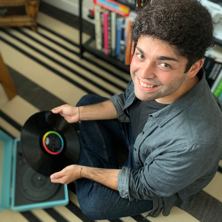 Raj looks up and smiles at a camera. He is holding a vinyl with a rainbow ribbon.