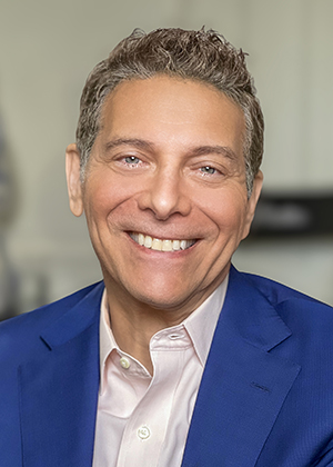 A man in a blue jacket and light collared shirt smiles.