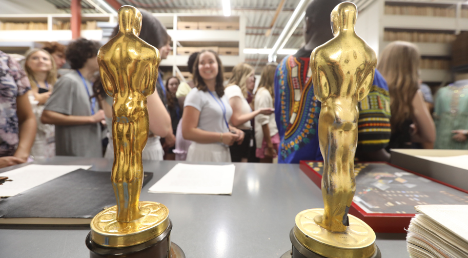 Two gold Academy Award statuettes in an Archive space with a group of high school age students smiling as they explore the artifacts.
