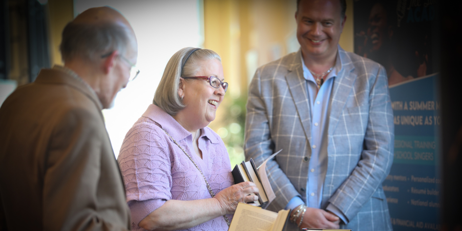 An older woman with bobbed blonde hair, glasses and a purple top holds books and smiles. Two men stand beside her and look at her.