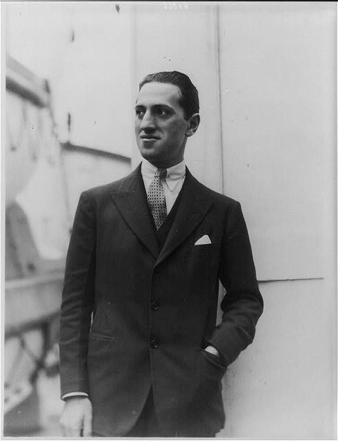 A black and white photograph of George Gershwin, a young man with dark hair slicked back, wearing a dark suit, vest, patterned tie, and a white pocket square. He is standing against a light-colored wall, looking slightly to the side with a gentle smile.