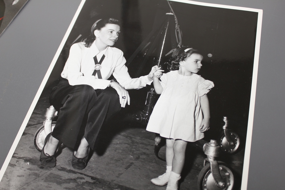 Judy Garland sits back stage and holds a young Liza Minnelli's hand. They both look to the right.