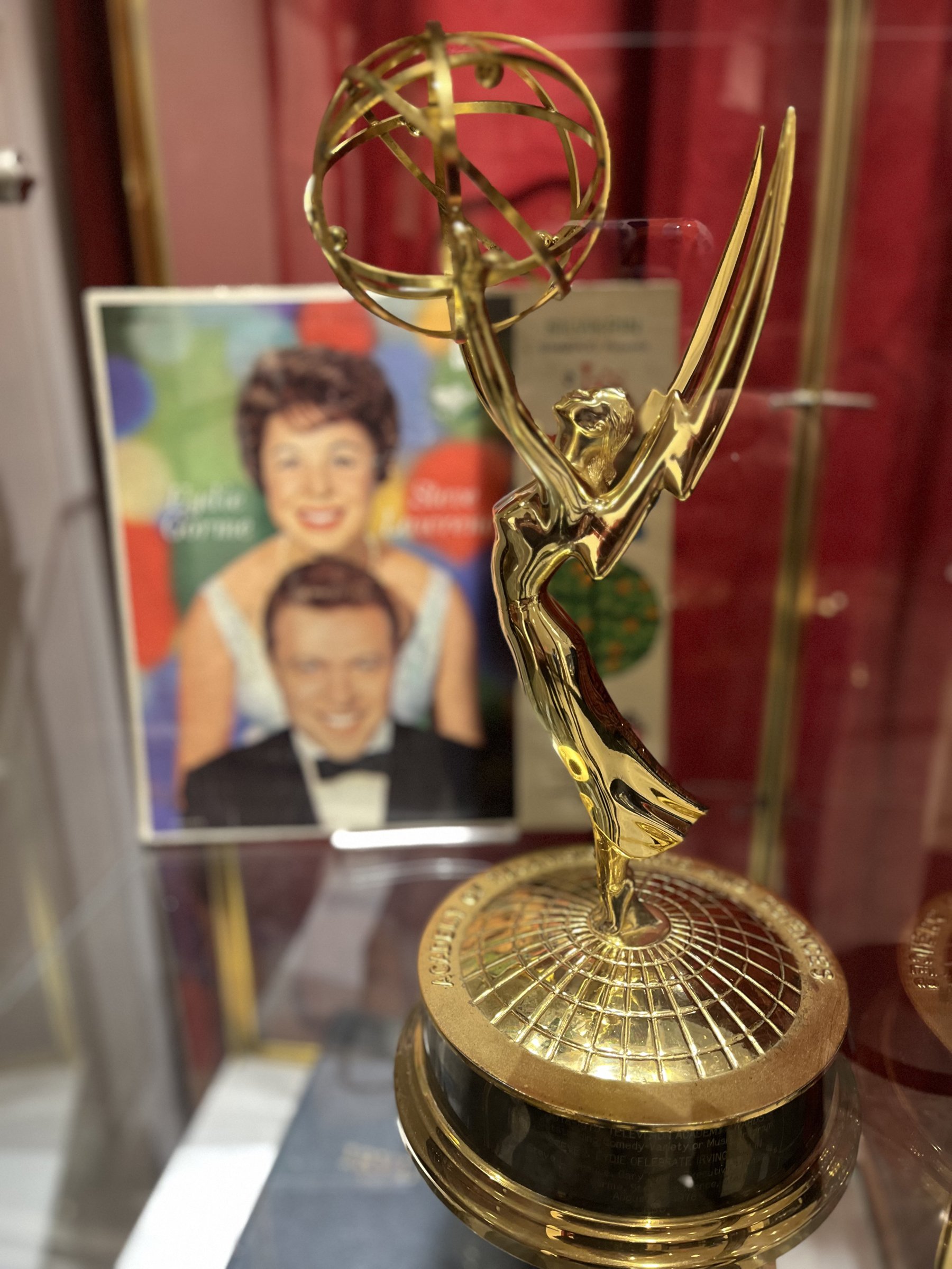 A shiny gold Emmy Award stands in front of a soft-focus photo of singers Eydie Gormé and Steve Lawrence.