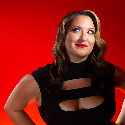 A woman in black dress, hands on hips, smiles in front of a red background