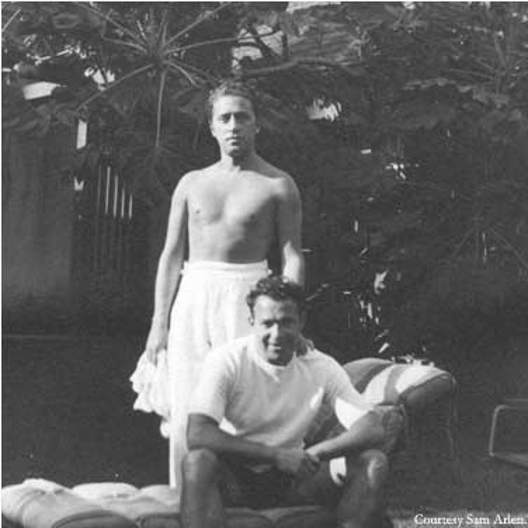 Harold Arlen stands in a towel with his hand on lyricist Yip Harburg's shoulder. Harold wears a solemn look while Yip wears a grin. They are in front of palm trees.