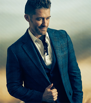 A man with short dark hair looks downward as he poses for a photo in a blue checked suit.