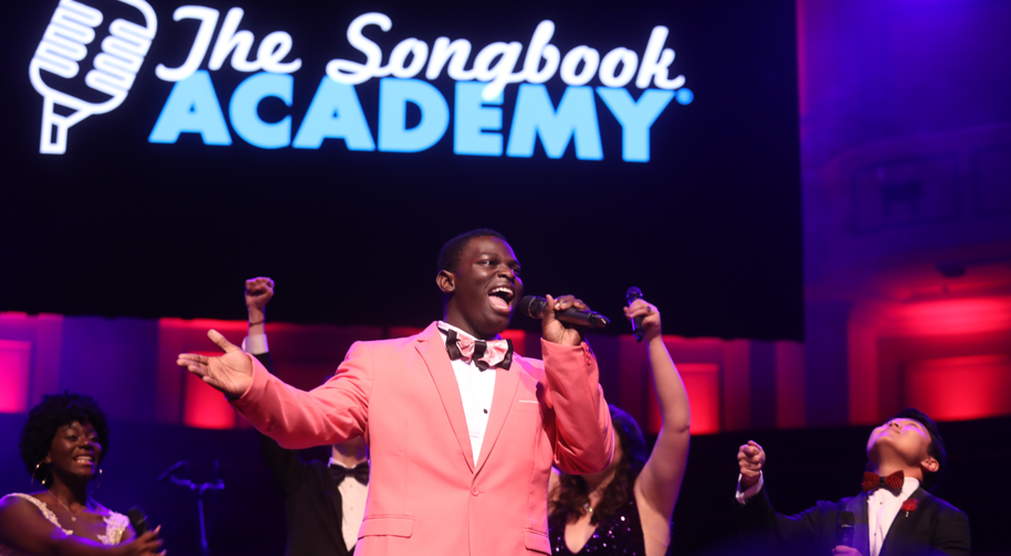 J'lan, a young Black singer in a coral suit holds a microphone and sings on stage. Behind him are other singers in various poses and the Songbook Academy logo above their heads.
