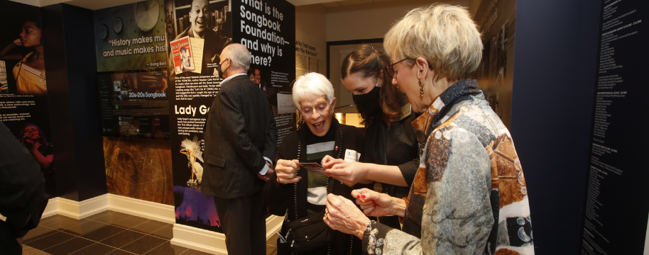 Two older women and a middle aged woman in a mask look at an object in the Songbook Exhibit Gallery.
