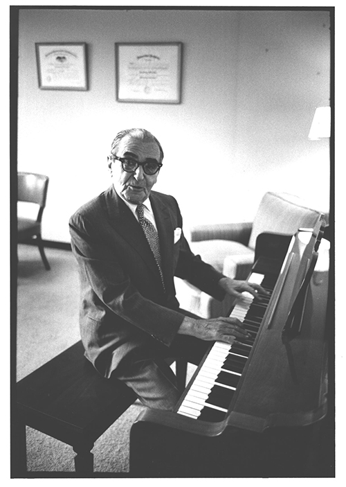 An elderly man in a suit and eyeglasses plays a piano.