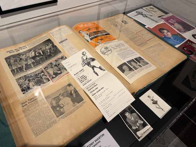 Scrapbook displayed in a museum case, opened to pages of vintage newspaper clippings, programs, and photos related to Cyd Charisse and Tony Martin. Additional portrait photographs and memorabilia are arranged around the album under glass.