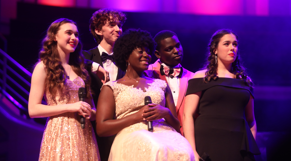 Five high school singers are posed as a group on a stage. They all smile and look to the left.