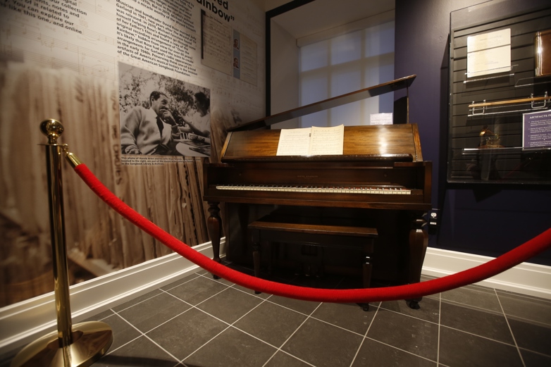 A dark wood upright piano with a large fallboard stands behind a red rope. Beside the piano is a photo of Harold Arlen and his wife.