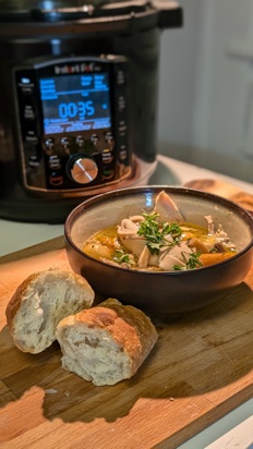 Image of a delicious-looking bowl of chicken and vegetables in broth. Crusty bread sits in the foreground and an Instant Pot in the background.