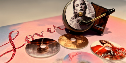 A music box with a crank displays an image of a woman. Around it are small discs with various portraits of the same woman in different poses, placed on a surface that reads "Lover" in sparkling red script.