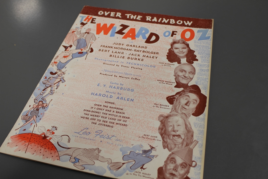Red, blue and sepia toned sheet music for "Over the Rainbow" from the movie musical "The Wizard of Oz". The pictures feature Judy Garland as Dorothy, the professor, the Scarcrow, the Cowardly Lion, the Tin Man and images from the original book. The sheet music also has names of songs featured in the film.