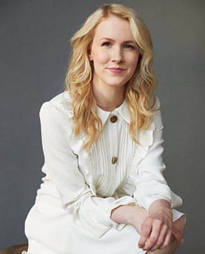 A blonde woman poses in a white dress, hands in her lap.