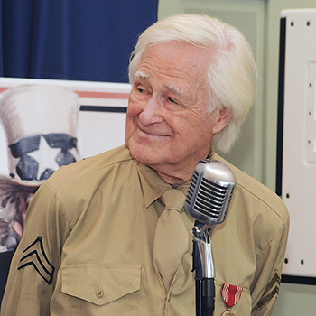 An elderly man in a brown military shirt stands at a microphone, with patriotic images in the background.
