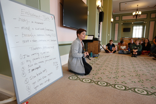 Broadway star Mary Michael Patterson sits on the carpeted floor beside a dry erase board in the center of students, also sitting on the floor. The board lists "Embodiment Tools" for mindfulness.