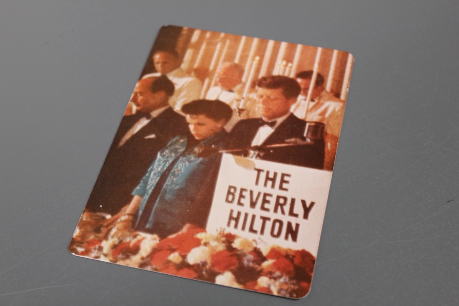 An older Judy Garland stands next to President John F. Kennedy in front of a sign that reads "The Beverly Hilton". They are dressed in cocktail attire and Judy looks down with a sullen look.