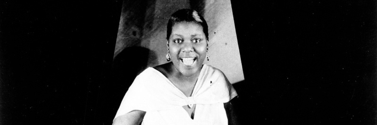 Bessie Smith wears a white dress with a cape and smiles broadly at the camera.