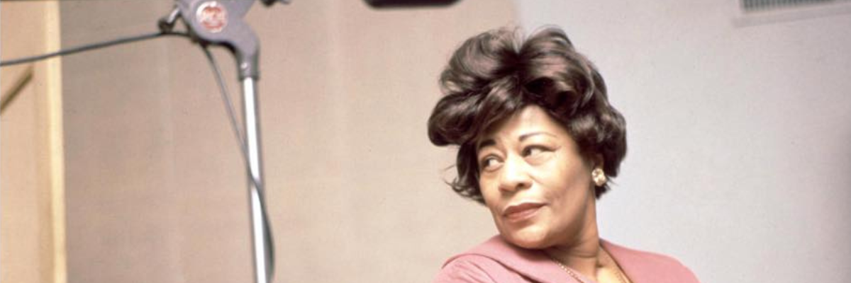 Ella Fitzgerald is in a recording studio and looks to the left. She is wearing a rose colored blouse and has a 60s hairdoo.