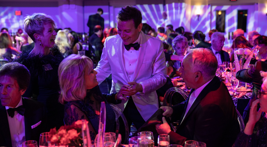 Michael Feinstein is dressed in a white tuxedo and talks to donors. They are in a purple lit concert hall at a gala.