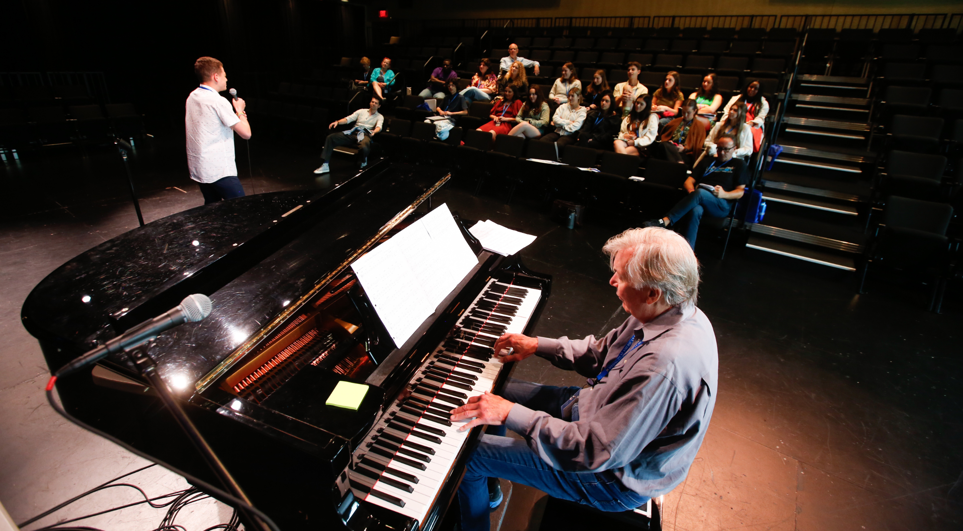 A pianist plays for a student singing into a microphone for a small audience.